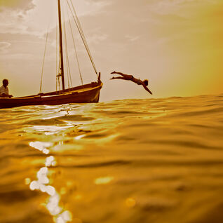 Sunset water level photo of a person diving off a fishing boat into the ocean with an orange sky